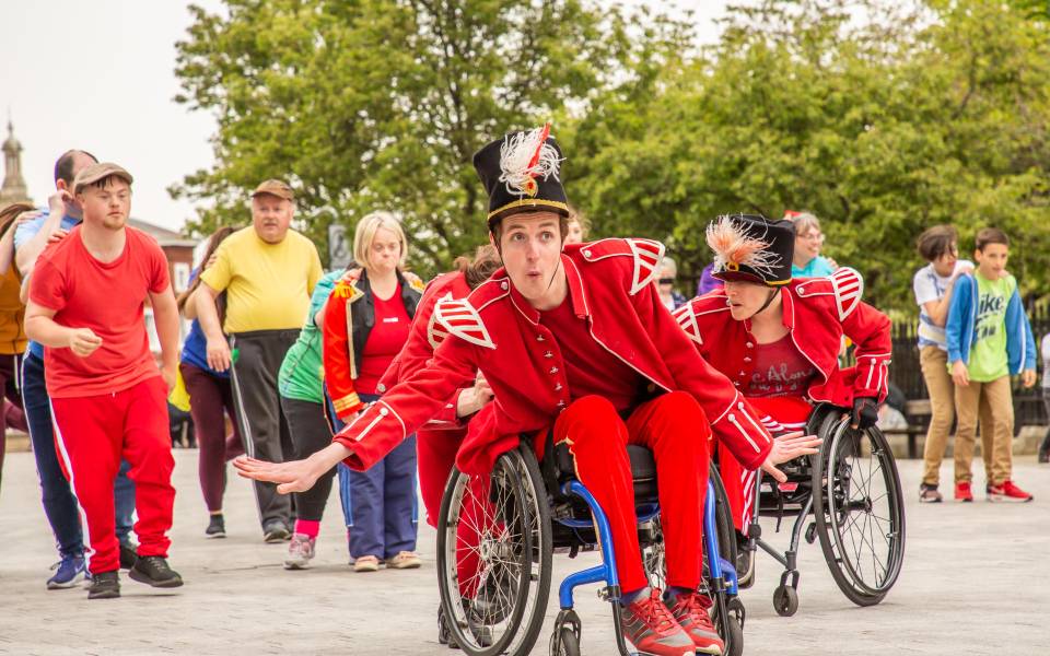 An outdoor performance of dance show Frippery. A man in a wheelchair, and wearing an all red marching band outfit and hat, has his arms outstretched. Behind him are more dancers wearing bight colours.