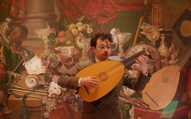 A man stands before an ornate painting playing a lute.